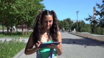 A beautiful girl in a green T-shirt with black hair walks through the park using a tablet. slow motion. 4K video