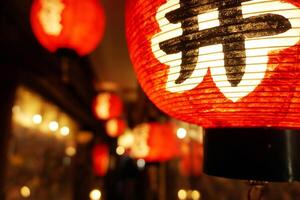 de cerca negro japonés textos en rojo papel linterna colgando debajo japonés restaurante alero en noche hora con borroso antecedentes. foto
