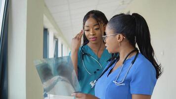 dos joven afroamericano hembra doctores en sinfh trajes examinar un radiografía y discutir eso video