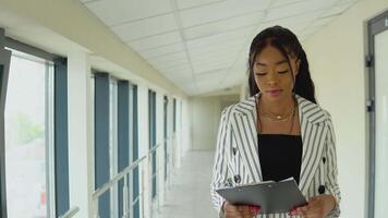 atractivo africano americano mujer de negocios en blanco a rayas traje en pie por el ventana en el pasillo de el oficina centrar video