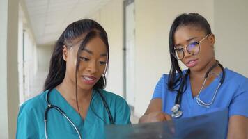 dos joven afroamericano hembra doctores en sinfh trajes examinar un radiografía y discutir eso video