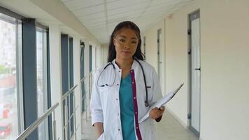 joven africano americano médico mujer en un azul traje y blanco Saco camina a lo largo el corredor de el clínica con documentos en su mano video