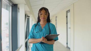 uma jovem africano americano fêmea médico dentro uma azul terno carrinhos dentro a corredor do a clínica e examina a documentos video