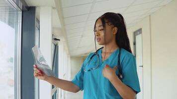 une Jeune Afro-américain femelle médecin dans une bleu costume des stands dans le couloir de le clinique et examine un radiographie de un adultes poitrine video