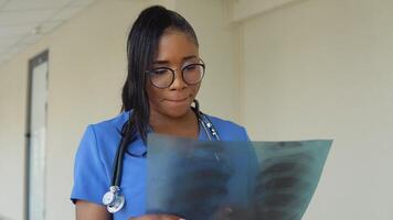 un' giovane afroamericano femmina medico nel un' blu completo da uomo sta nel il corridoio di il clinica e esamina un raggi X di un di adulto il petto. video