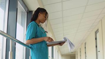 jovem africano americano médico mulher dentro azul terno carrinhos dentro clínica corredor e vira através documentos video