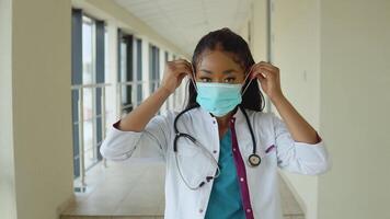 Jeune africain américain médecin femme dans une bleu costume et blanc manteau porte une jetable stérile visage masque video