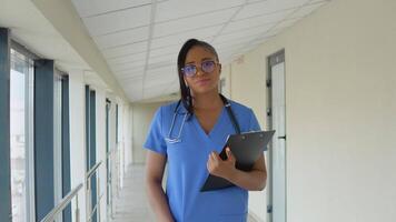 joven africano americano médico mujer en un azul traje camina a lo largo el corredor de el clínica con documentos en su mano video