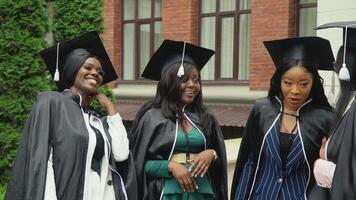 feliz universidade ou Faculdade graduados dentro animado mantos e quadrado chapéus comunicar animado e alegremente. afro-americano fêmea alunos ficar de pé perto a universidade construção video