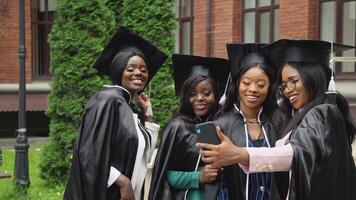 gelukkig Universiteit of college afgestudeerden in meester jurken en plein hoeden zijn levendig en nemen selfies. Afro-Amerikaans vrouw studenten staan in de buurt de Universiteit gebouw video