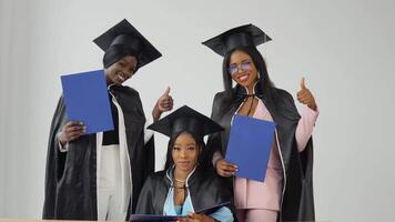One woman is sitting at a desk, and two classmates are standing behind her and smiling. Happy graduates of a university or college of African American nationality with blue diplomas in their hands video