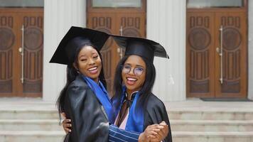 Two African-American female graduates in classic master's suits and robes stand embracing in front of the university entrance. Happy graduates of the student exchange program. video