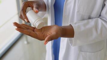 africano americano médico mujer en un azul traje y blanco Saco vierte pastillas desde un tarro en su palmera. de cerca ver de manos video