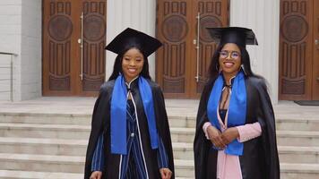 dos afroamericano mujer graduados en clásico maestría trajes y batas estar en frente de el Universidad entrada. contento graduados de el estudiante intercambiar programa video