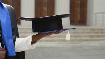 An African-American graduate woman in a black master's gown proudly and cheerfully holds a diploma and a square hat against the backdrop of the university. An important event. Close view of a hand video