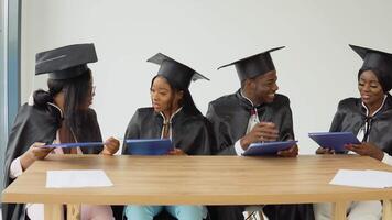 vier afgestudeerden van een Universiteit of college van Afrikaanse Amerikaans nationaliteit zitten met diploma's in hun handen Bij een bureau en hebben pret pratend naar elk andere video
