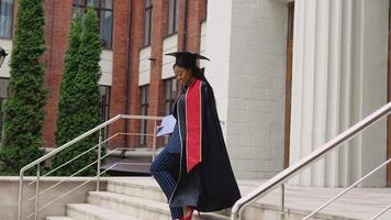 un afroamericano hembra graduado en un negro túnica y un maestría sombrero con orgullo y alegremente camina arriba el escalera a el Entrada a el Universidad con un diploma de mayor educación en mano video