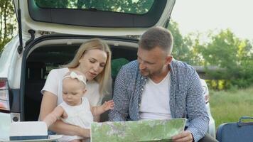 gelukkig familie op reis door auto. een vader en moeder houden een baby meisje in hun armen en kijken Bij een weg kaart. actief vrije tijd voor de familie video