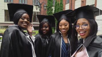 gelukkig Universiteit of college afgestudeerden in meester jurken en plein hoeden zijn levendig en nemen selfies. Afro-Amerikaans vrouw studenten staan in de buurt de Universiteit gebouw video