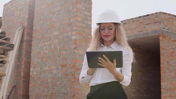 kvinna arkitekt i företag klädsel och en vit hjälm står i en nytt byggd hus med obehandlad väggar och Arbetar på en läsplatta. modern teknik i de äldsta yrken video