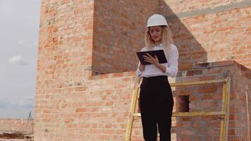kvinna arkitekt i företag klädsel och en vit hjälm står i en nytt byggd hus med obehandlad väggar och Arbetar på en läsplatta. modern teknik i de äldsta yrken video