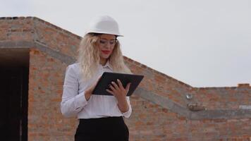 vrouw architect in bedrijf kleding staat in een nieuw gebouwd huis met onbehandeld muren en werken Aan een tablet. modern technologieën in de oudste beroepen video