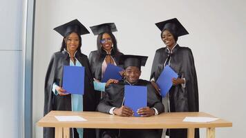 une homme est séance à une bureau, et Trois femmes sont permanent derrière lui. diplômés de une Université ou Université de africain américain nationalité avec bleu diplômes dans leur mains video