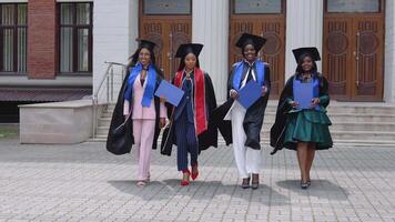 contento laureati di un' Università o Università di africano americano nazionalità con blu diplomi nel loro mani camminare inoltrare per il telecamera a partire dal il Ingresso per il Università video