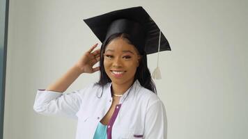 een jong Afrikaanse Amerikaans vrouw afstuderen in een meester hoed en wit medisch japon glimlacht poses voor de camera. een belangrijk evenement. jong specialist video