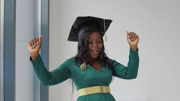 An African American female graduate in a green holiday dress with gold accessories and a square master's hat stands with a diploma in her hands and dances. Higher education for women abroad video
