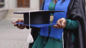 An African-American graduate woman in a black master's gown holds a diploma and a square hat against the backdrop of the university. An important event. Close view of a hand video