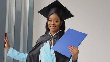 un africano americano hembra graduado en un clásico maestría traje y manto soportes por el ventana con un diploma en su manos y sonrisas un graduado de Universidad con un azul diploma en su manos video