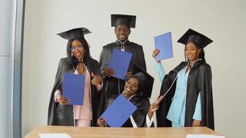 One woman is sitting at a desk, and three classmates are standing behind her. Happy graduates of a university or college of African American nationality with blue diplomas in their hands video