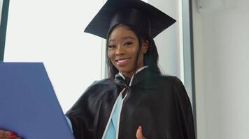 un africano americano hembra graduado en un clásico maestría traje y manto soportes con un diploma en su manos y sonrisas un graduado de un médico Universidad con un azul diploma en su manos video