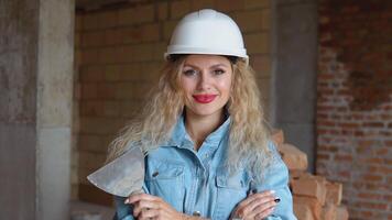 A female builder in a helmet and denim work uniform stands on a construction site. Young woman bricklayer with makeup and manicure holding a spatula video