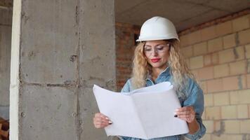 bouwkundig bouw plan. een jong vrouw in een wit werk helm en denim overall en stofbril staat Bij de bouw plaats en houdt een bouw plan. video