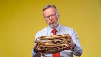 Frustrated executive worker over yellow background overworked holding piles of documents paper work. photo