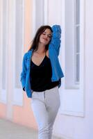 Girl posing in light hall. Hand over head, closed eyes. Casual clothes. White jeans, black top and blue shirt. Blurred background. photo