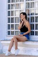 Young attractive girl sitting on windowsill near the wooden window. Touching face with hand. Black top and blue jeans. photo