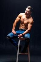Man with perfect body sitting on high chair. Dark background. Male beauty. photo