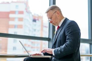 Side view on working manager. man in suit with laptop near big window. photo