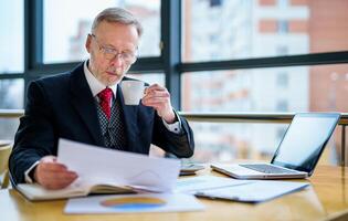 Elderly businessman using laptop and taking notes working while sitting in modern office. Selective Focus photo