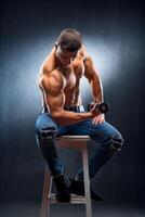 Man with perfect body pumping up muscles with barbell in hand. Athlete sitting on high chair. photo