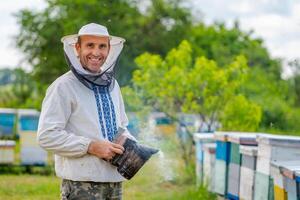 Beekeeper at apiary at the summer day. Man working in apiary with beesmoker. Apiculture. Beekeeping concept. photo