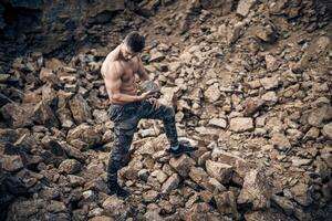 Full size photo of an athletic guy posing with rock in a quarry. Outdoor shot. Bodybuilder. Quarry background. Closeup.