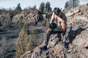 Full length photo of a bodybuilder with metal dumbbell in hand. Shirtless bodybuilder. Rocks on the background.