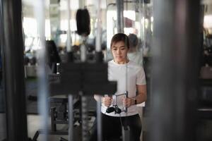 asiático deporte trabajando fuera a aptitud gimnasia, levantamiento pesos con un barra con pesas. ella es enfocado y determinado, probable molesto a mejorar su fuerza y aptitud física. el gimnasio es equipado con varios máquinas. foto