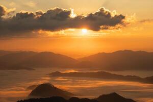 The sun is setting over a mountain range, casting a warm glow over the landscape. The sky is filled with clouds, creating a sense of mystery and depth. The mountains are covered in fog photo