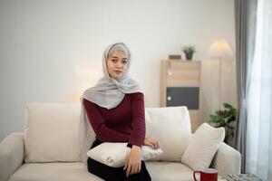 Muslim Islam woman wearing hijab or grey scarf sits on a couch with a white pillow. She is looking at the camera photo