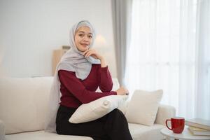 Muslim Islam woman wearing hijab or grey scarf sits on a couch with a white pillow. She is smiling and she is relaxed. A cup and a book are placed on a table nearby photo